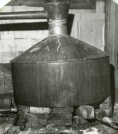 Stacks of brinks in a circle support a metal moonshine kettle with a pipe coming out the top at a right angle.