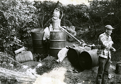 A man in a plaid shirt stands in a wooded area with over turned barrels and piping. Bags and other refuse are scattered about.