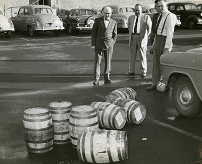 3 white men in suits stand in a parking lot looking at 7 barrels of liquor. 