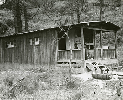 A cabin built of worn planks in a forest setting. A man stands on the porch and a broken liquor barrel lays in the font on grass