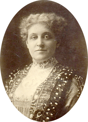 Photo of Carrie Chapman in a fancy lace & embroidered dress. Her light hair is parted down the middle & tied back.
