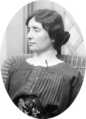 Photo of Helen Keller sitting in a wicker chair, wearing a dark dress with high white collar. Her hair is parted in the middle and tied behind with a wide bow.