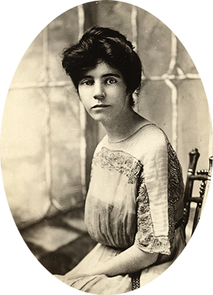 Photo of Alice Paul sitting in a wood straight back chair & wearing a dress with embroidery.