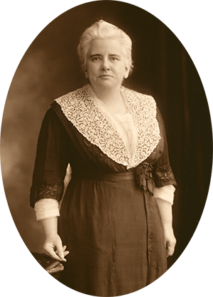 Photo of Anna Shaw standing next to a table. She wears a black dress with lace collar. Her hair is white with age.