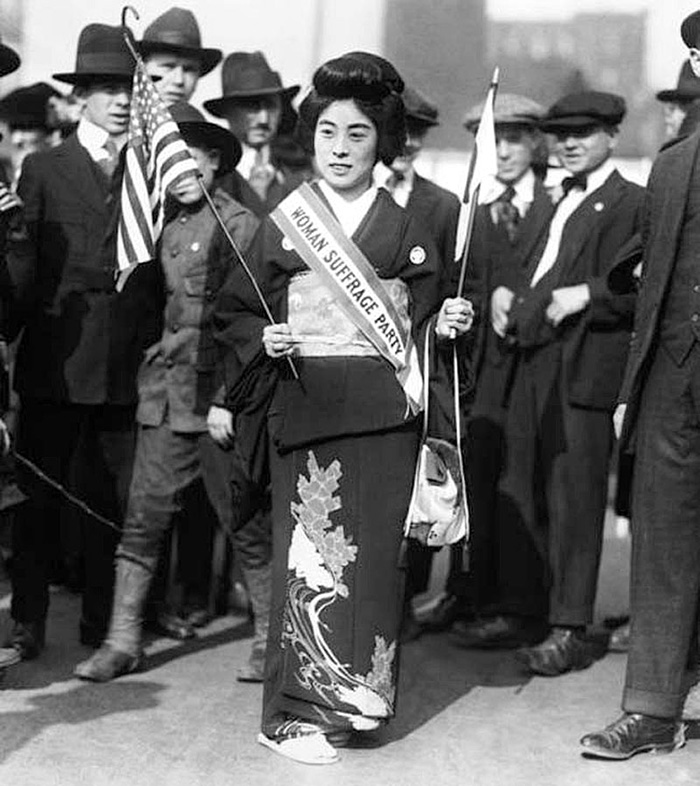 Japanese woman in kimono with women suffrage sash. Men stand behind her.