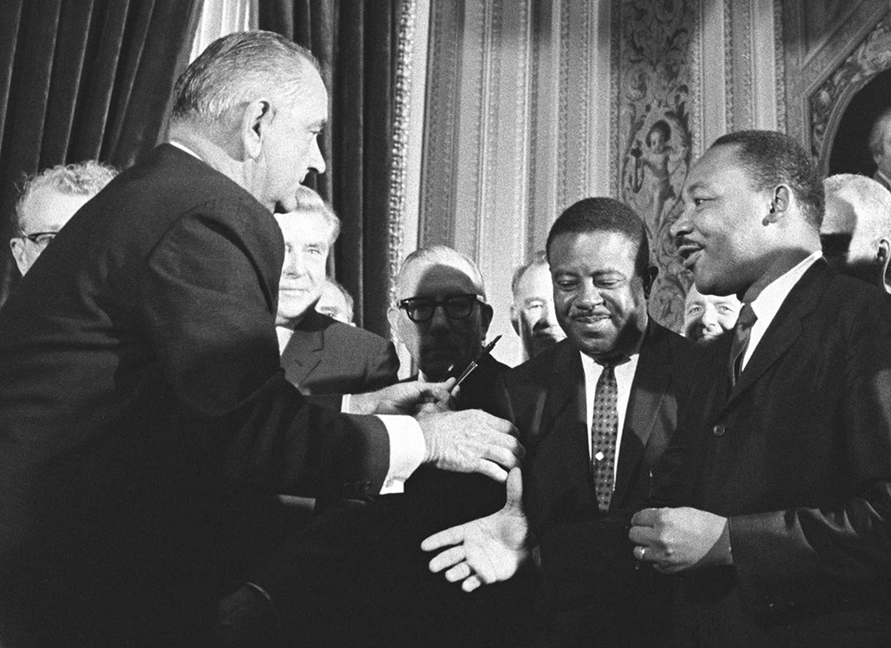Lyndon B. Johnson and Martin Luther King shake hands while surrounded by a group of men.