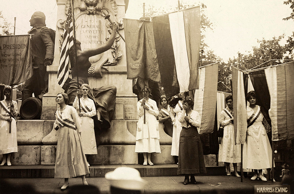About 10 women with tall banners stand of the steps of a momunment to General LaFayette.