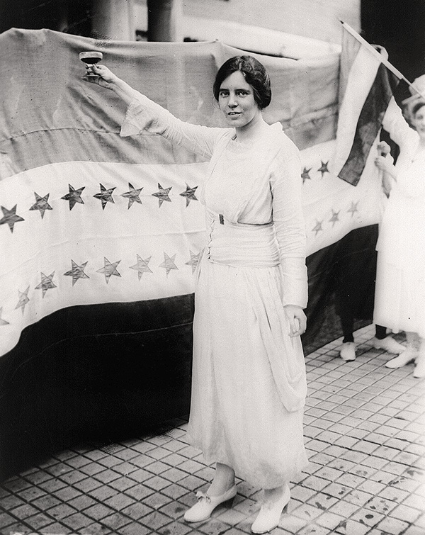 Photo of Alice Paul standing before a banner with a glass of liquid raised in her right hand.