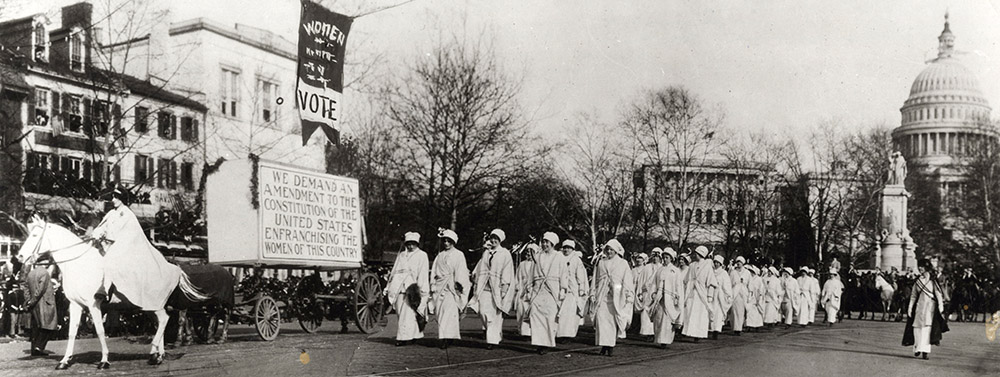 Photograph from 1913. Woman on white horse leads a formation of approximately 50 women through a street. The capitol building is in the background.