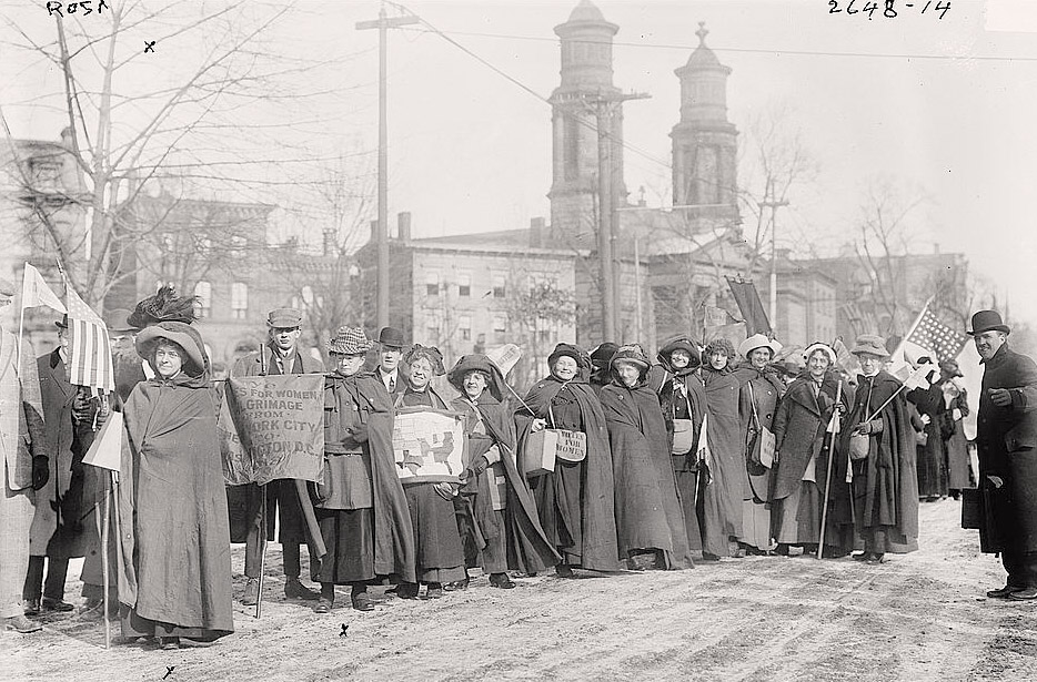 a line of women, and a few men, dressed for cold weather stand on a snow dusted street waving flags and signs for suffrage.