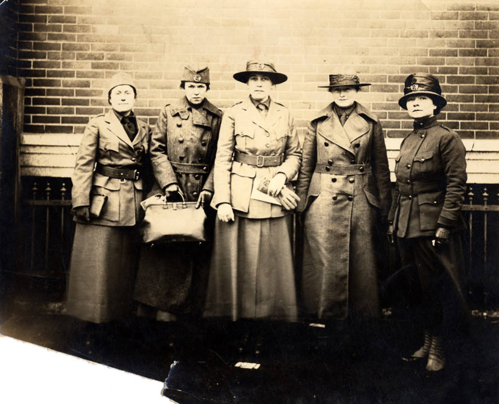 Photo of 5 women dressed in military service type clothing standing before a brick wall.