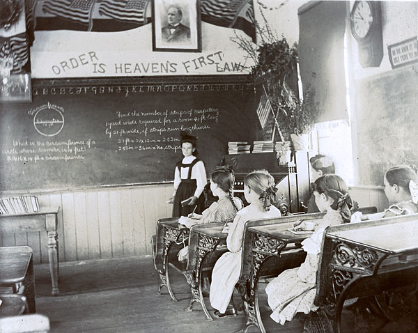 Teacher stands at blackboard. Above her on wall reads: Order is Heavens First Law