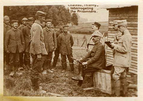 German prisoners stand on left as four men in uniforms question them. One of the 4 men sits on a crate holding a cane.