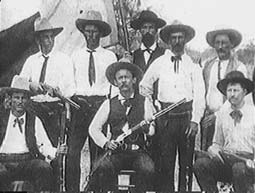 Black and white photo of 8 men in cowboy hats and brandishing guns.