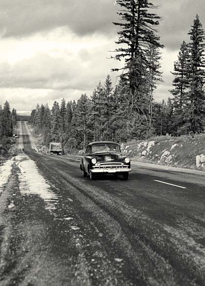 A car of the time period travels down a 2 lane road lined with evergreen trees. It looks like there might be remains of snow.