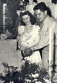 Woman in wedding dress, man in military uniform cutting a wedding cake.