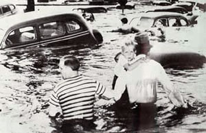 2 men, one holding a child, wade through water up to their waists. Cars flooded up to their windows stand in their way.