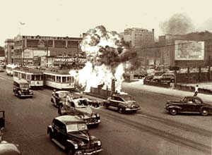 Photo of something on fire in the middle of a city street in Detroit. Cars and street cars are close to the fire in the street.