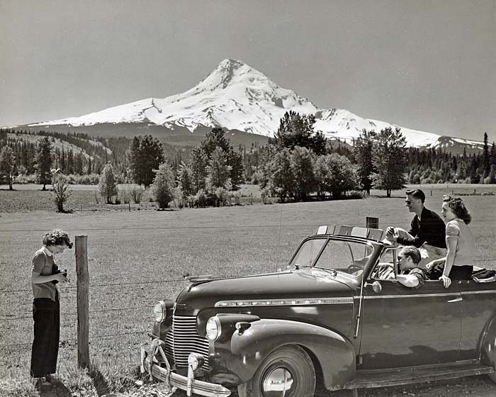 2 men & woman in their 20's pose for a photo in their car as another woman with a camera stands outside the car. Mt. Hood behind