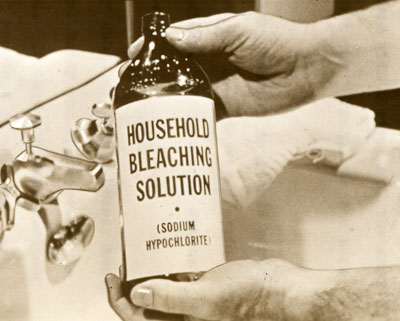 2 hands hold a bottom over a sink. The bottle reads: Household Bleaching Solution.