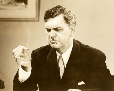 A man in a suit and tie holds a lit cigarette in his right hand and looks at it with a frown on his face and furrowed brows.
