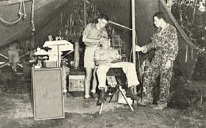 2 men standing, 1 man sitting in a chair inside a tent in the forest. 