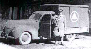 Man in military uniform stands next to 1939 Ford ambulance with door held open.