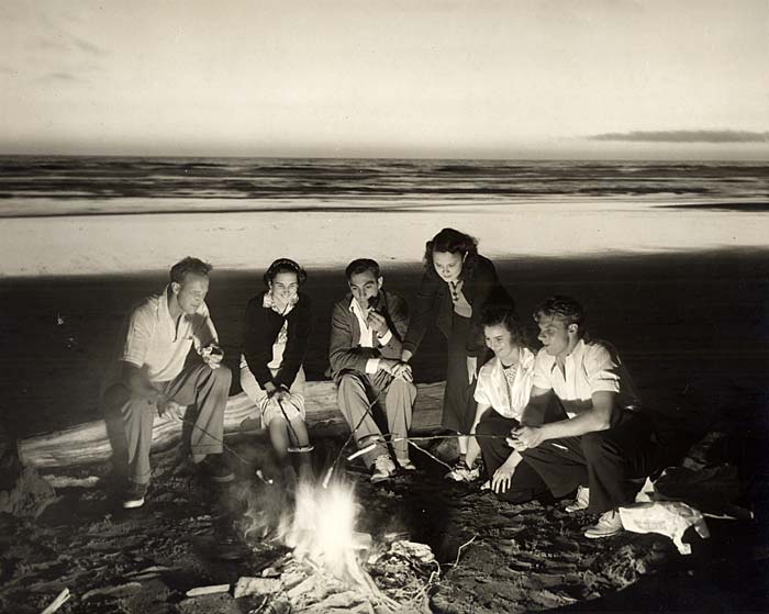 6 people huddle around a camp fire on the beach. They sit on logs and hold sticks with hotdogs on the end into the fire.