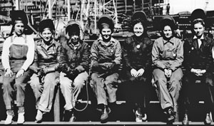 7 women in welders helmets sit on a curb or sidewall. 