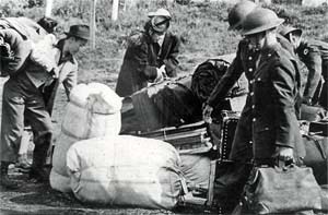 2 Japanese Americans pick up bundles with their belongings as men in military uniforms assist.