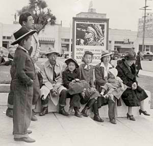 About 6 or 7 Japanese American people sit on a curb waiting. They include men, women, children and one infant.