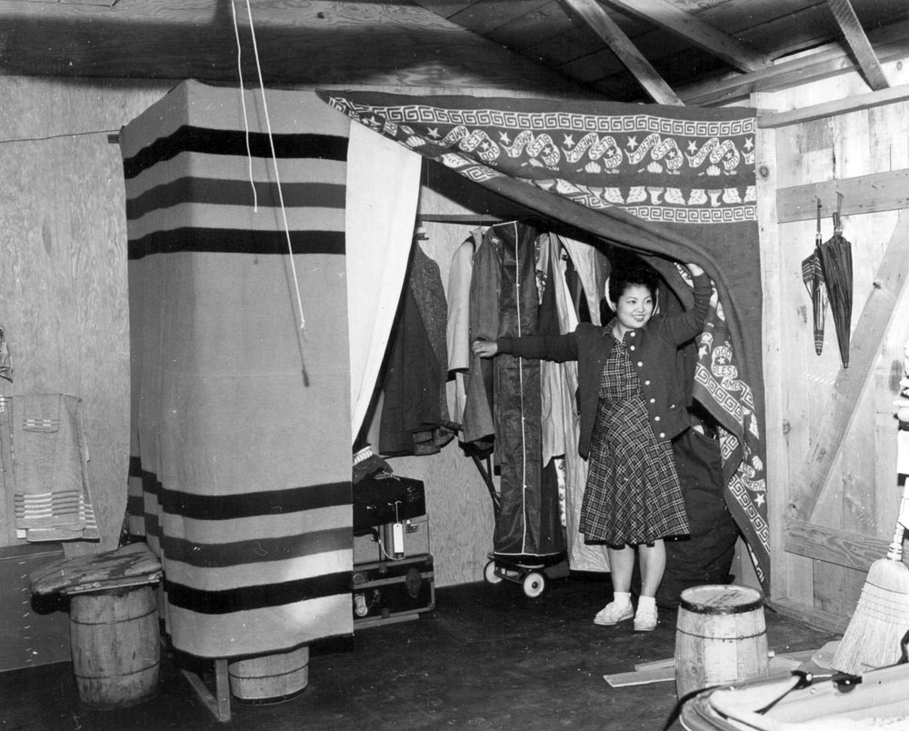 A Japanese American woman pulls back a currtain that has been hung about 8 ft off the floor as a makeshift closet. Inside are clothes hanging.