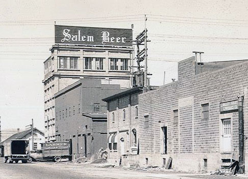 Brick building with sign on top says Salem Beer. Trucks from the 1930s shown on street.