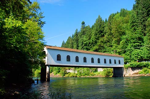 Covered bridge with windows in side wall spans a river. Thick forest stands on the bank of each side.