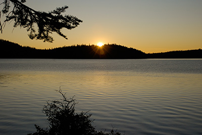 Calm lake with banks cloaked in darkness the sun peeks our from behind a hill as it sets.