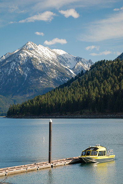 Boat docked at a walkway with snow capped mountain in background. The near shore is covered in evergreen trees.