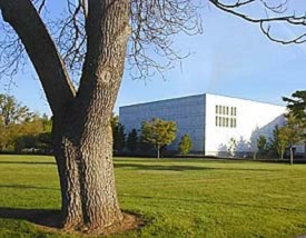 Square, white, windowless building on a lawn of green grass with small trees around.