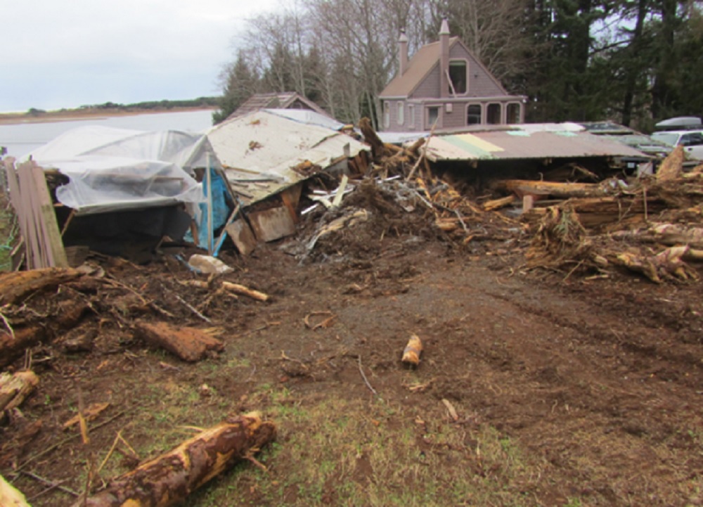 Landslide shows a mess of logs and debris with a house still standing in the background.