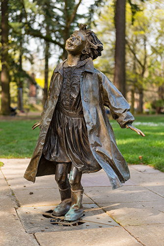 A bronze life‑size statue of Ramona Quimby, a spirited young girl in a raincoat and boots, poised mid‑step in a playful splash fountain.