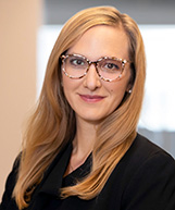 Christina Stephenson has long blonde hair and is wearing glassess and a black suit.