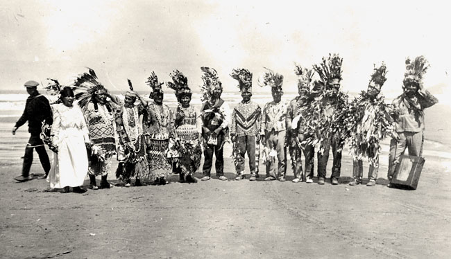 A dozen people standing in a line on a beach dressed in Native American clothing.