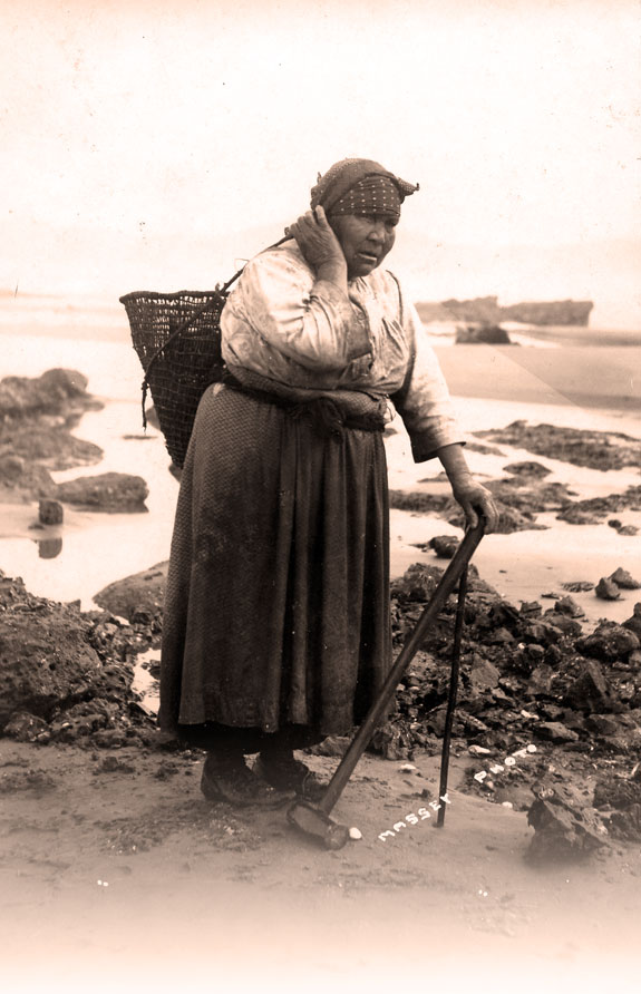 Native American woman with basket on her back. She's standing on a rocky shore with a sledge hammer & prybar in her hand.