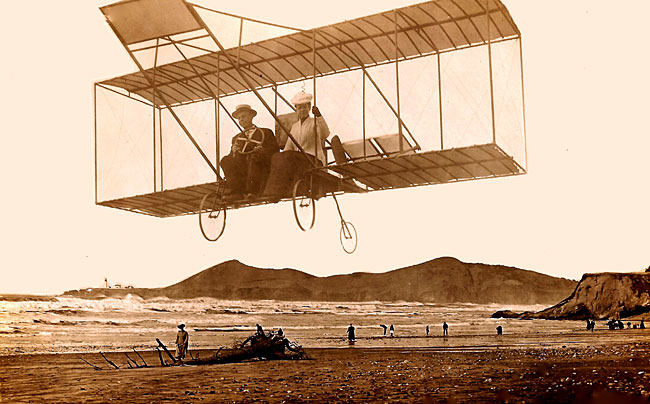 2 people on a glider style aircraft flying over a beach in 1911 with the ocean in background.