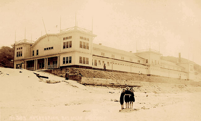 1914 photo of 3 people standing on a sandy beach in fron of the building Bayocean Natatorium.