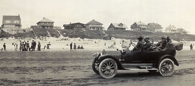 A model T type car on the hard sand of a beach with 4 people riding inside. Houses line a hill in the background.