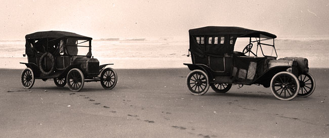2 model T Ford cars sit on the hard sand in front of the ocean.