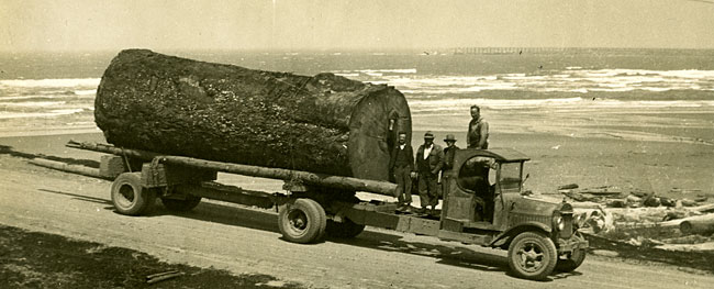 A 1920s log truck with a huge old-growth log on the back. 4 men stand on the bed of the truck, behind the cab. Ocean behind.