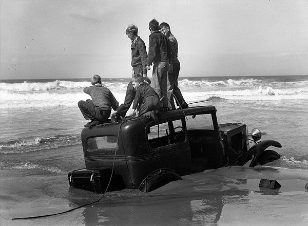 A 1920s style car sunk up to the tops of the tires in the sand at the ocean edge. 6 boys play on the car.