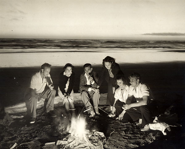 Black & white photo of 6 you adults around a campfire on the beach. They sit on a fallen log. It is dusk or evening. 