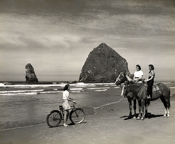 Photo of a woman with a bicycle, 2 women on horses on the beach. Haystack Rock is in the background.